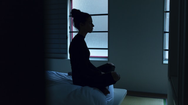 Een vrouw zit in een donkere kamer op haar bed en staart in het niets, omdat haar man kanker heeft.